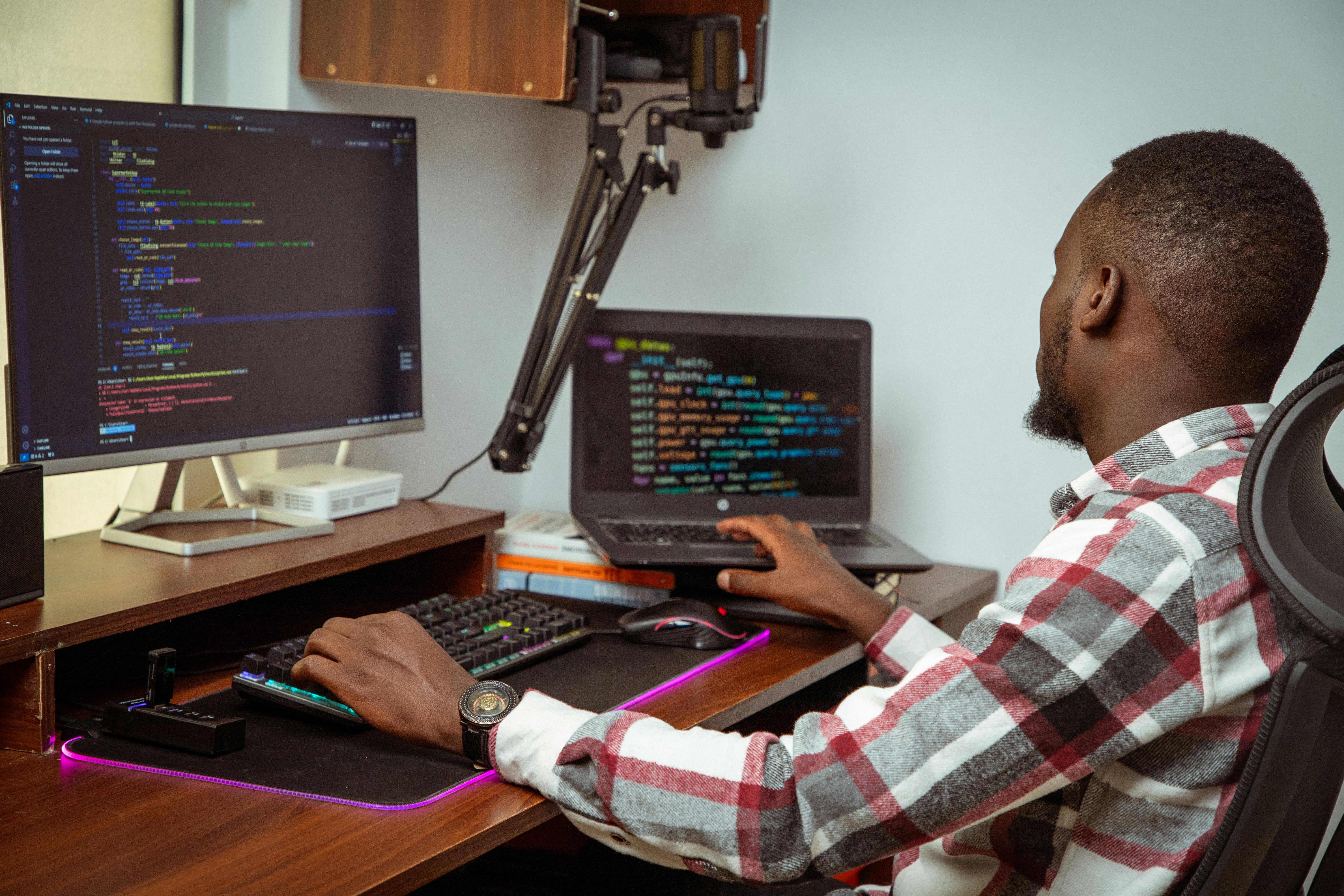 An African man coding on a desktop and laptop in a Nairobi office setting, showcasing modern technology.